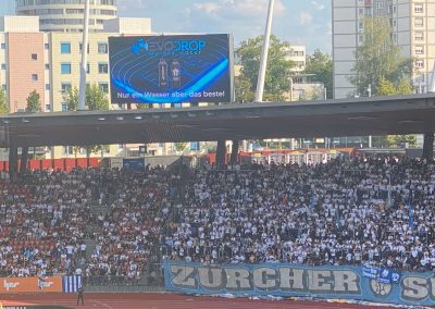 Grossbildschirm im Zürcher Fussballstadion mit Evodrop-Werbung und der Botschaft "Nur ein Wasser, aber das Beste!" über der jubelnden Zürcher Südkurve.